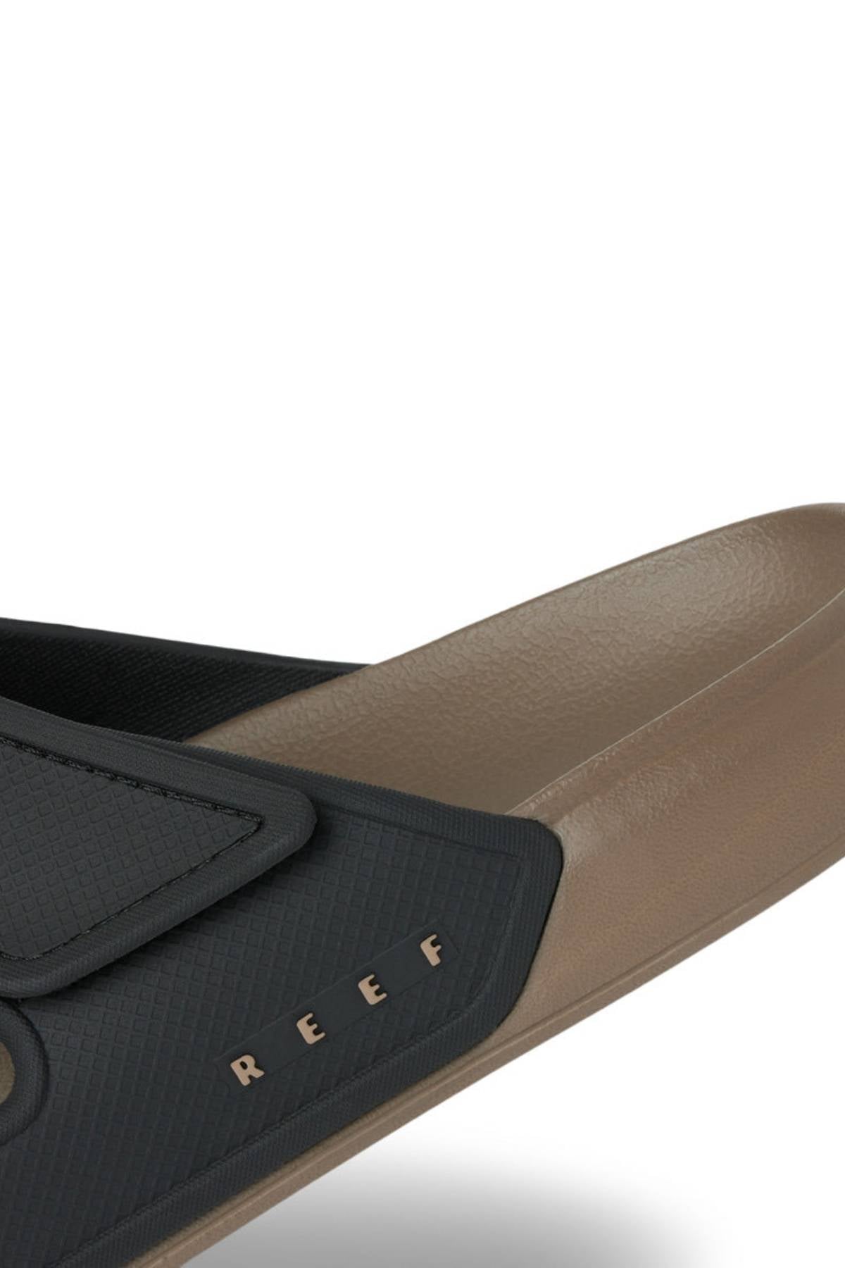 Close-up of the REEF Oasis Adapt sandal, showcasing its textured black strap with gold "REEF" lettering, comfortable EVA footbed, and brown sole, all set against a plain white background.