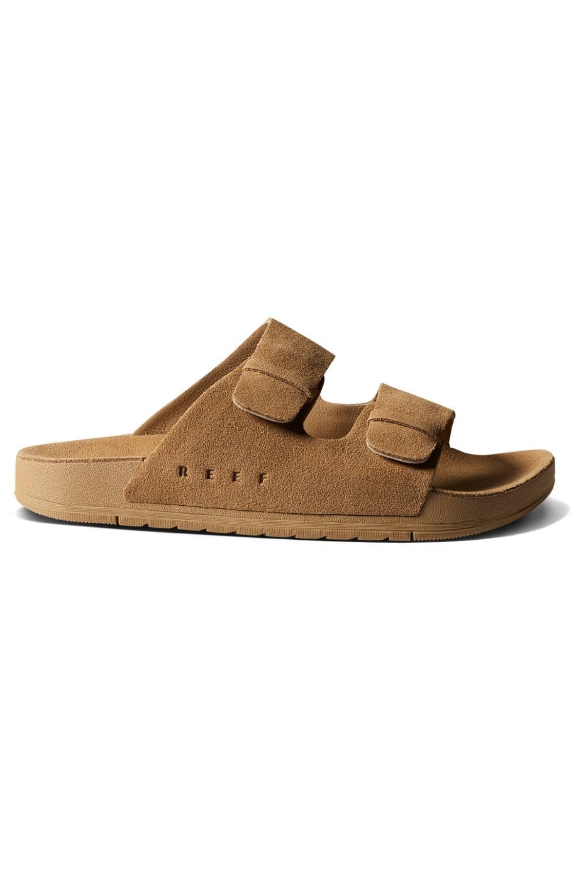 The REEF Ojai Two Bar is a tan suede slide sandal with two adjustable velcro straps, a flat sole, and "REEF" embossed near the heel, offering comfortable support ideal for all-day wear.