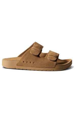 The REEF Ojai Two Bar is a tan suede slide sandal with two adjustable velcro straps, a flat sole, and "REEF" embossed near the heel, offering comfortable support ideal for all-day wear.