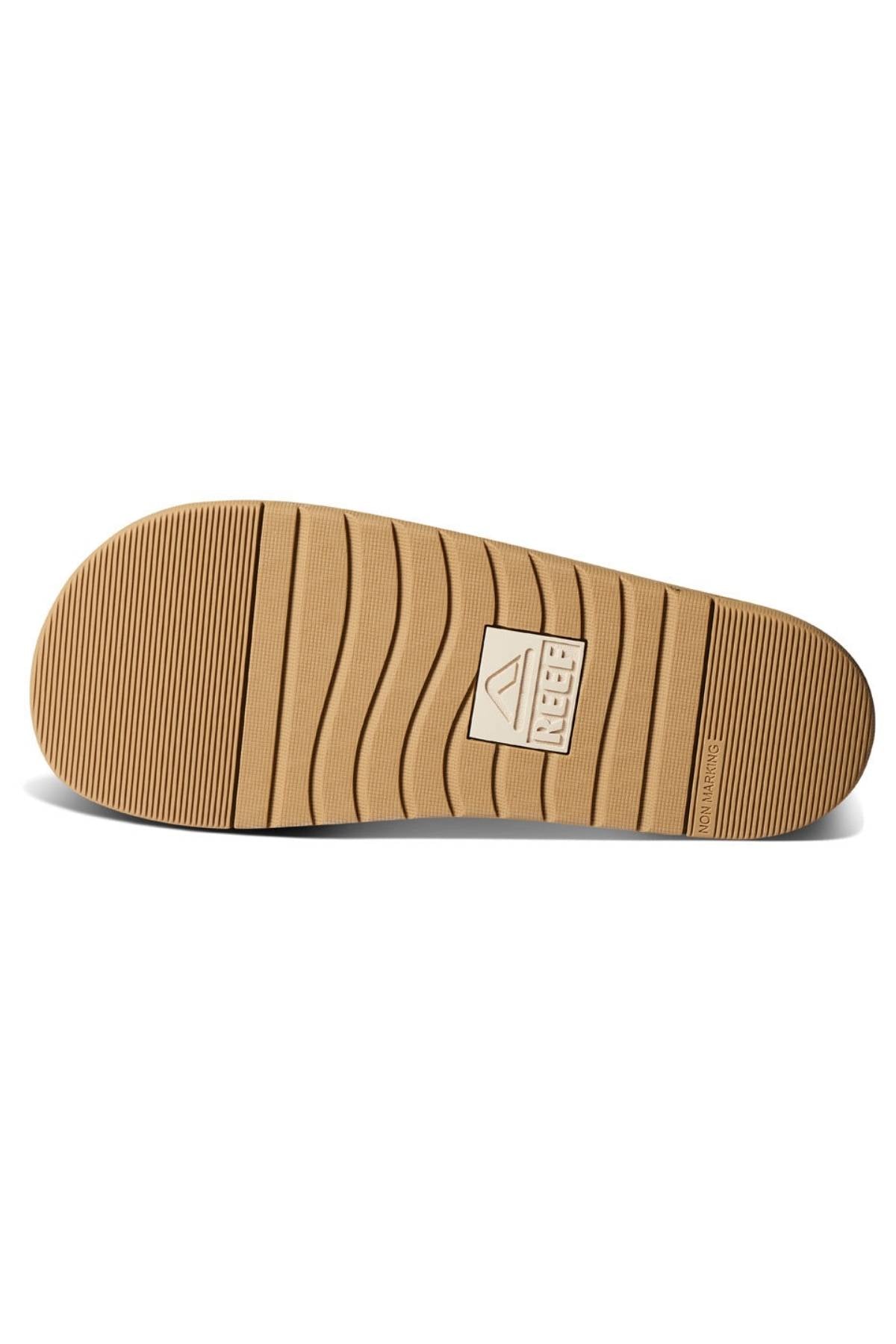 The image displays the bottom sole of the REEF Ojai Two Bar sandal in tan, featuring a ribbed tread pattern and a rectangular logo at the center, emphasizing the comfortable support typical of eco-friendly footwear.