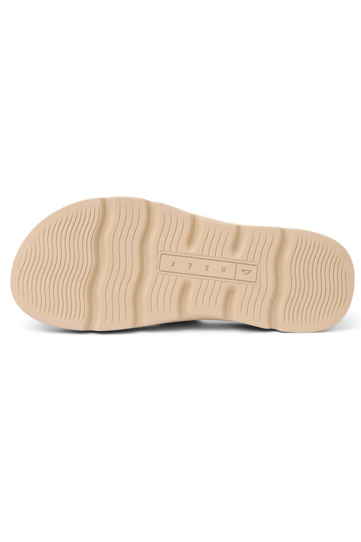 A beige rubber sole with wavy textured grooves for traction, designed for the REEF Vista Luxe Thea sandals, features a central rectangular embossed section with small symbols and raised lines for detail.