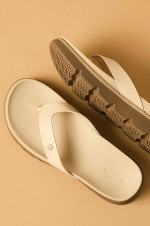 Two REEF Vista Luxe Vivianahh vegan leather sandals in cream, featuring thick, textured brown soles, are shown on a beige surface—one sandal lies flat, while the other is angled to display its tread pattern and cushioned footbed.
