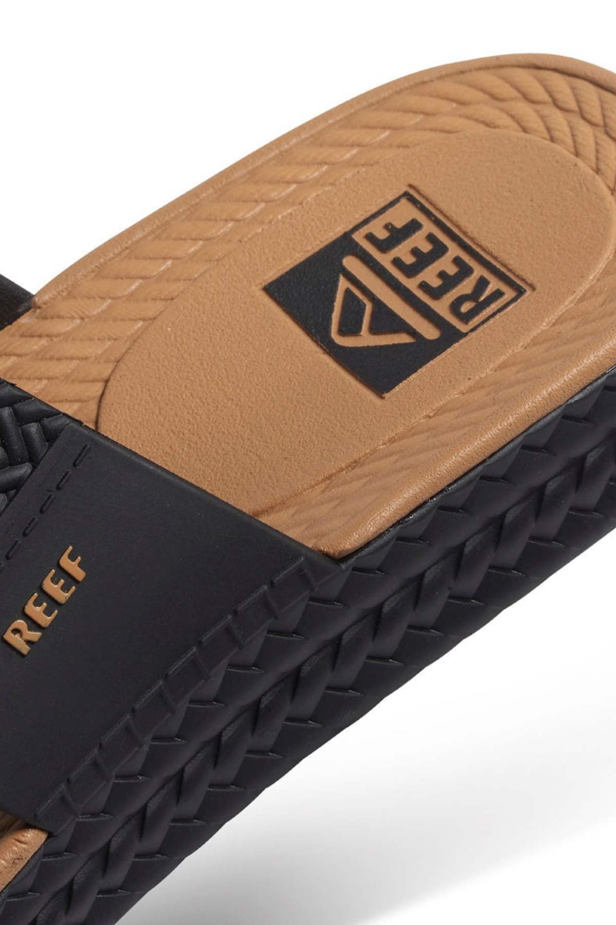 A close-up of the REEF Water Vista Slide sandal highlights its woven black strap with a basket-like design and "REEF" in gold. The tan dual-density EVA footbed, smooth with a rectangular logo, offers comfort and style, making it perfectly waterproof for all adventures.