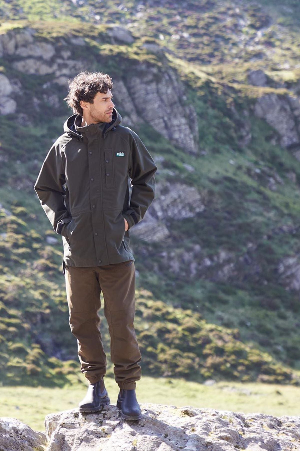 A man stands on a rock outdoors, wearing the Ridgeline Monsoon Classic Jacket in olive green, paired with brown pants and black boots. Behind him, green grassy hillsides and rocky terrain create a rugged natural backdrop.