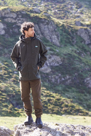A man stands on a rock outdoors, wearing the Ridgeline Monsoon Classic Jacket in olive green, paired with brown pants and black boots. Behind him, green grassy hillsides and rocky terrain create a rugged natural backdrop.