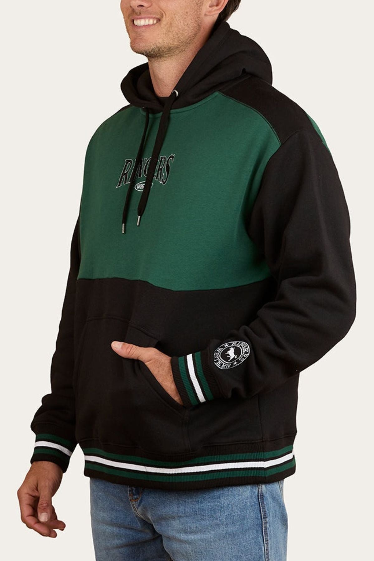A man smiles in a relaxed pose, wearing an oversized Ringers Western Barrow Creek Pullover Hoodie with “RANGERS” text and chest logo, featuring striped cuffs and a pouch pocket.