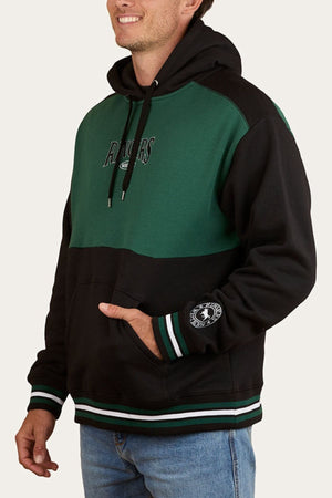 A man smiles in a relaxed pose, wearing an oversized Ringers Western Barrow Creek Pullover Hoodie with “RANGERS” text and chest logo, featuring striped cuffs and a pouch pocket.