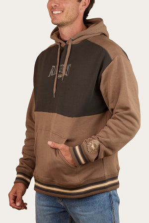 A man smiles in the Ringers Western Barrow Creek Pullover Hoodie—a brown and black color-blocked style with "Riders" on the chest, striped cuffs and hem—paired with blue jeans, standing against a plain light background.