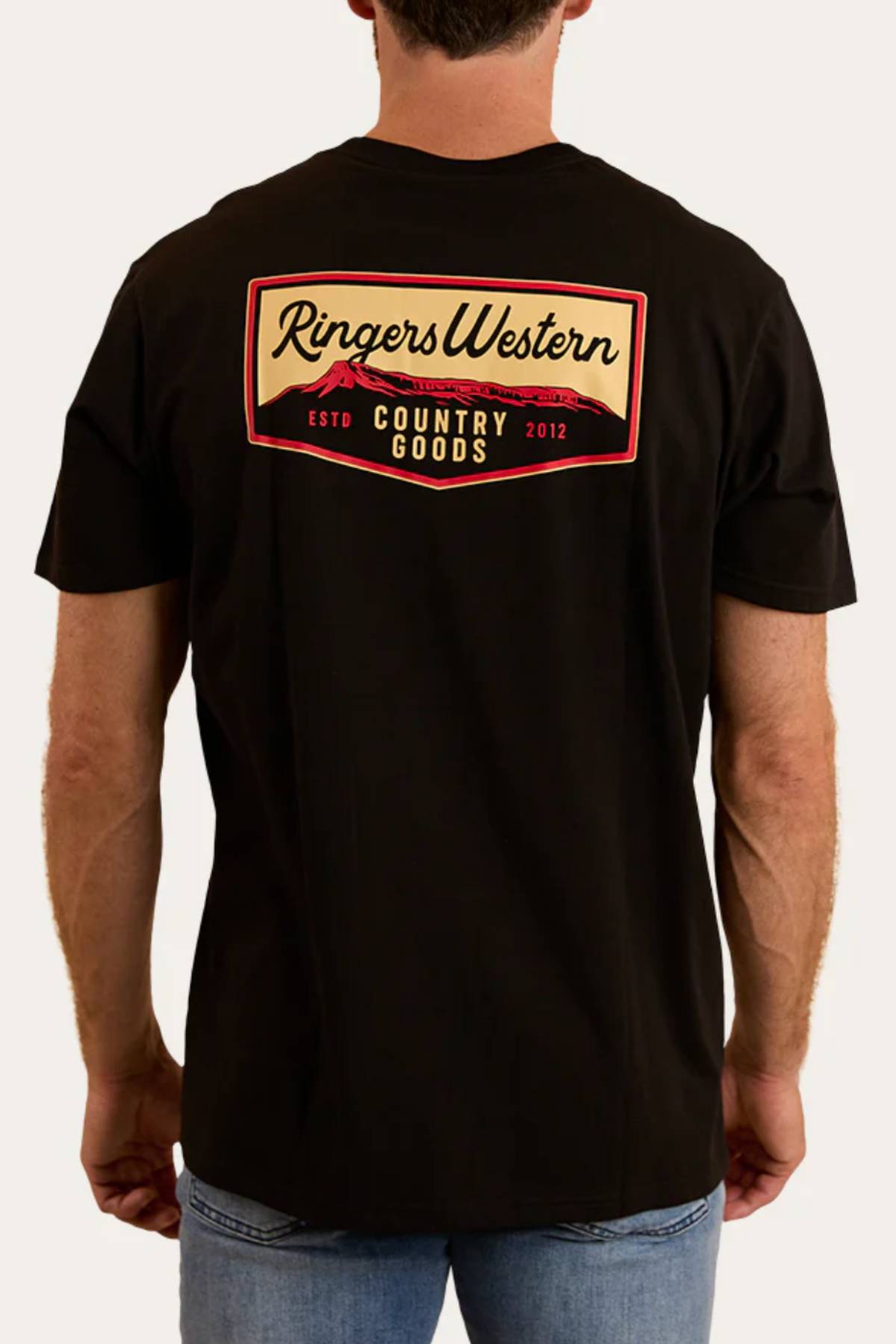 A man, facing away from the camera, wears a Ringers Western Country Goods Loose Fit T-Shirt with "Country Goods Estd 2012" in a red and yellow rectangle on the back, paired with light blue jeans.