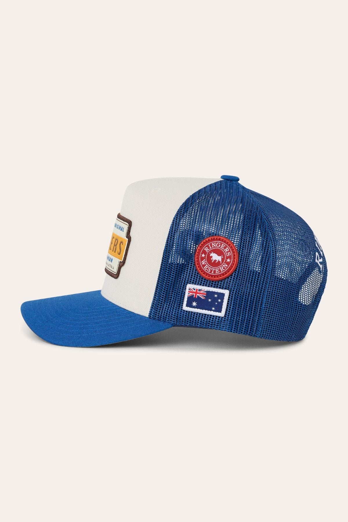 The Ringers Western Drive By Deep Fit Trucker Cap is a blue and white trucker with a deep fit and mesh back, adorned with multiple embroidered patches on the side—including a red circular patch, a rectangular Australian flag patch, and a partially visible front patch. Combining classic trucker styling with comfortable sun protection, this cap is perfect for everyday wear.
