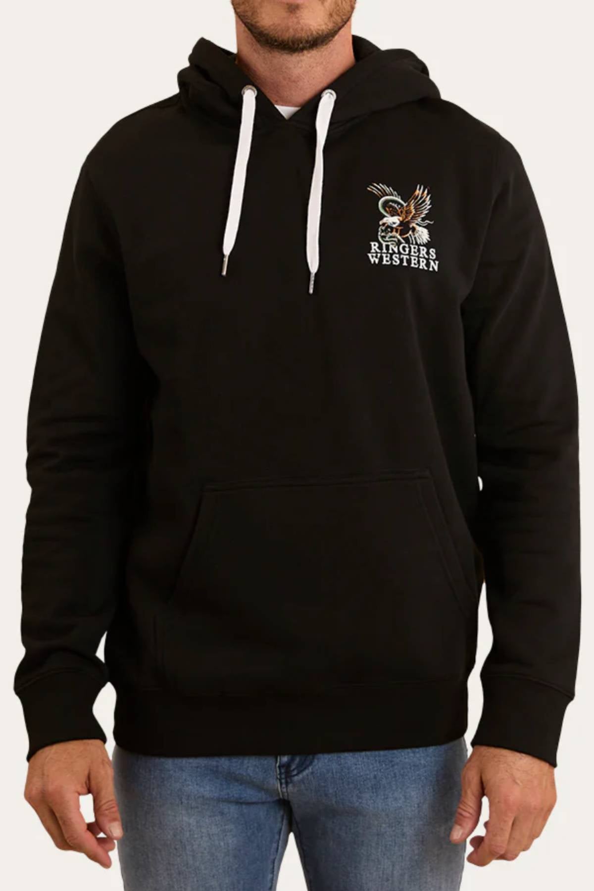 A man wears the Ringers Western Eyrie Pullover Hoodie—relaxed fit, black, with white drawstrings, a front pocket, and a small eagle graphic plus "Ringers Western" on the left chest—paired with blue jeans.