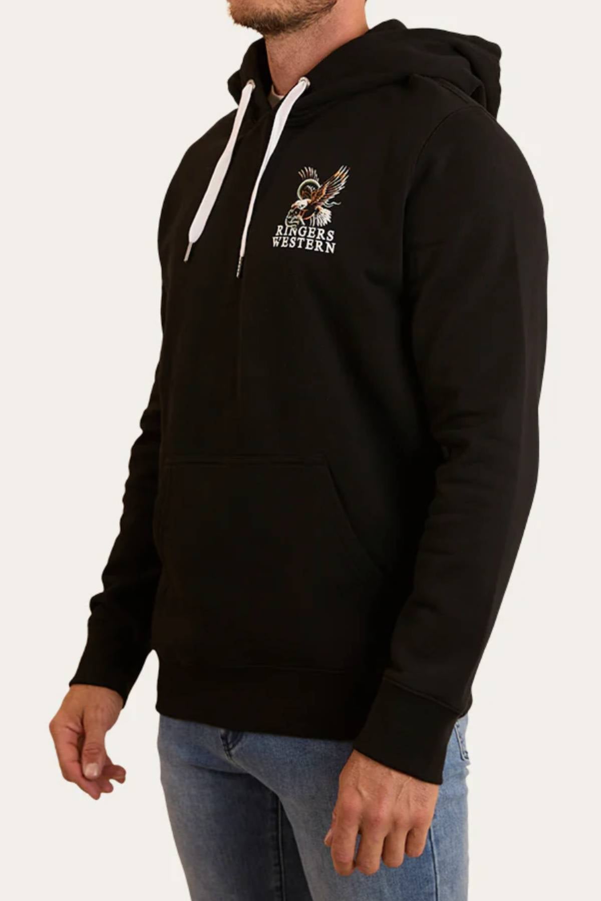 A man wears the Ringers Western Eyrie Pullover Hoodie—black brushed fleece with white drawstrings, front pocket, and a small "Ringers Western" eagle graphic on the left chest—paired with relaxed fit blue jeans.