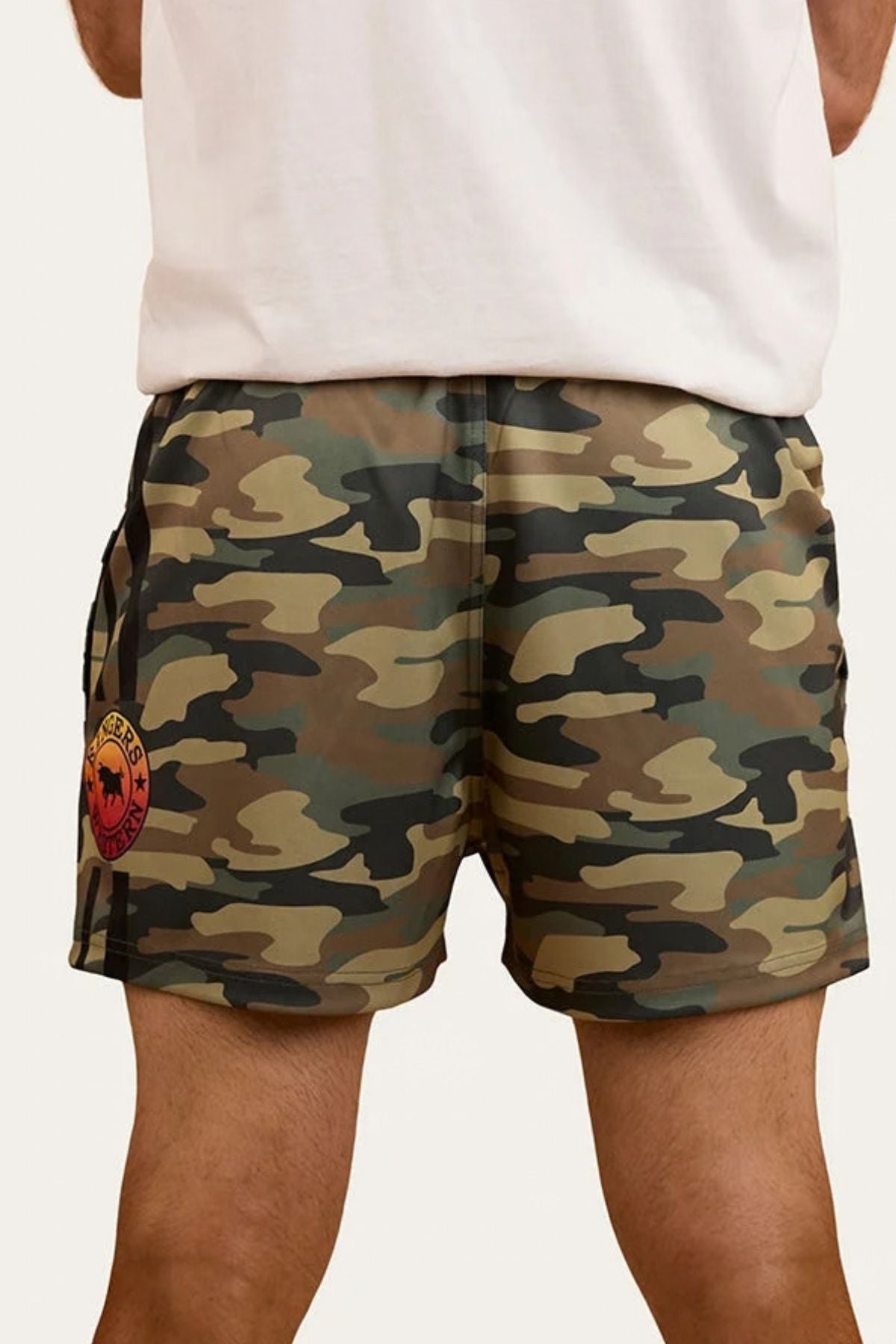 A person standing with their back to the camera is wearing Ringers Western Footy Shorts in green camouflage, featuring a high thigh cut and a red and yellow patch on the left side.