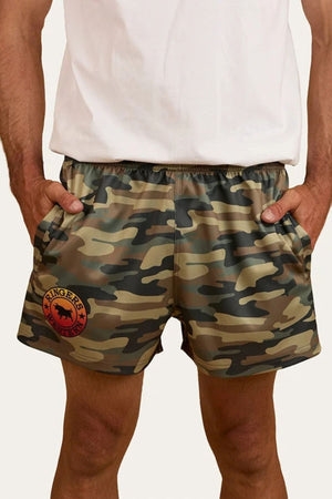 A person wearing a white shirt and Ringers Western Footy Shorts in camouflage, featuring a high thigh cut and red and black logo on the left leg, with their hands tucked into the shorts’ zippered side pockets.
