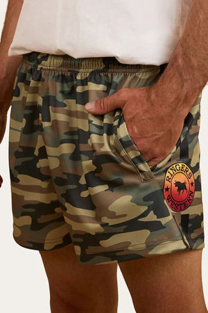 A person wearing Ringers Western Footy Shorts in camouflage with a red “Ringers Western” logo patch on the left leg, high thigh cut, and a hand tucked into one of the zippered side pockets.