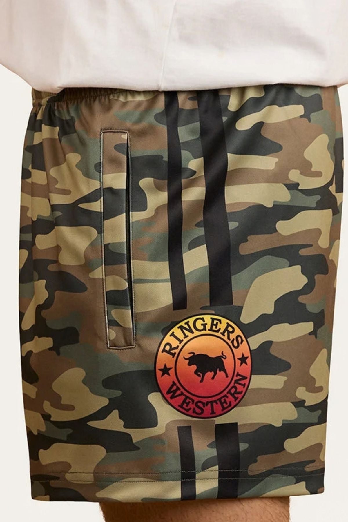 A person wearing Ringers Western Footy Shorts in green camouflage, featuring a black vertical stripe, a round “Ringers Western” bull logo, and zipper side pockets.