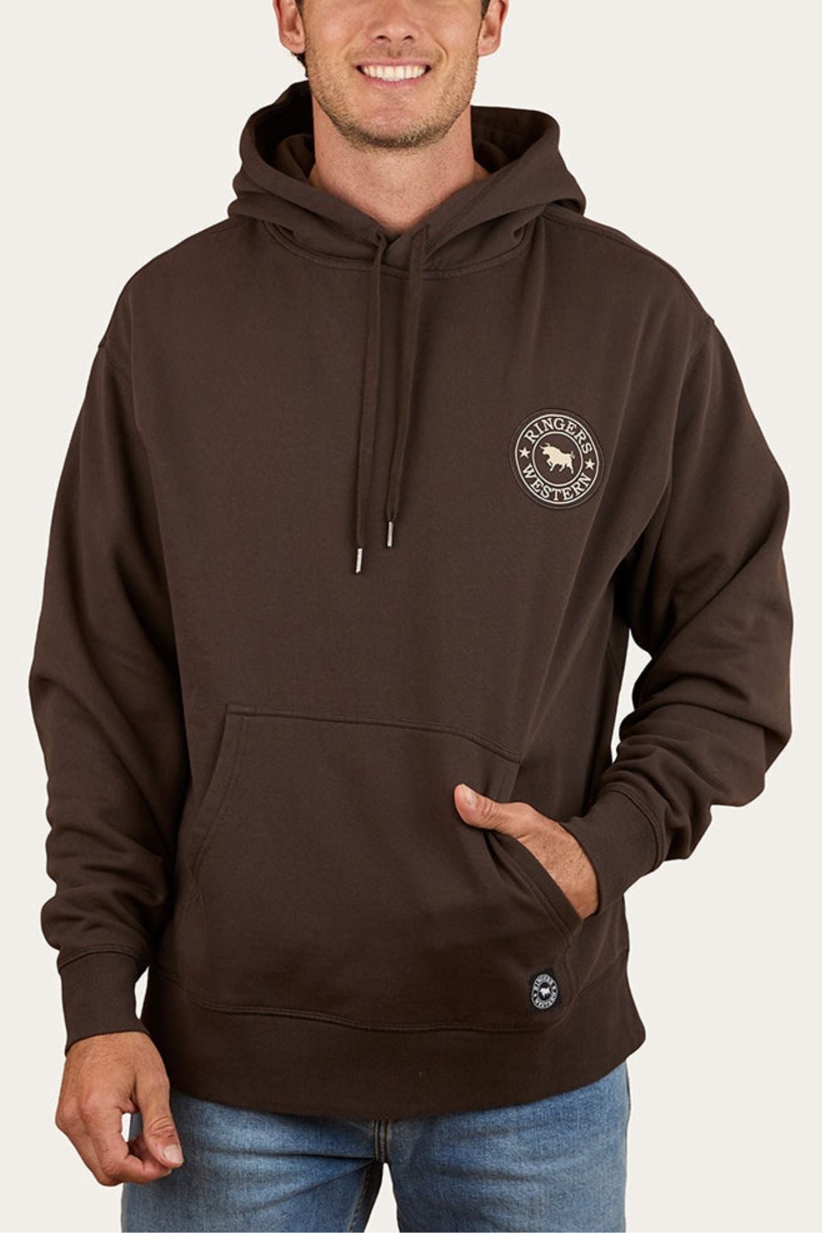 A man wears the dark brown Ringers Western Get Your Truck On Pullover Hoodie, featuring a front pocket, small circular left chest logo, drawstrings, premium brushed fleece, and a relaxed fit. He pairs it with blue jeans.