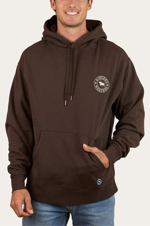 A man wears the dark brown Ringers Western Get Your Truck On Pullover Hoodie, featuring a front pocket, small circular left chest logo, drawstrings, premium brushed fleece, and a relaxed fit. He pairs it with blue jeans.