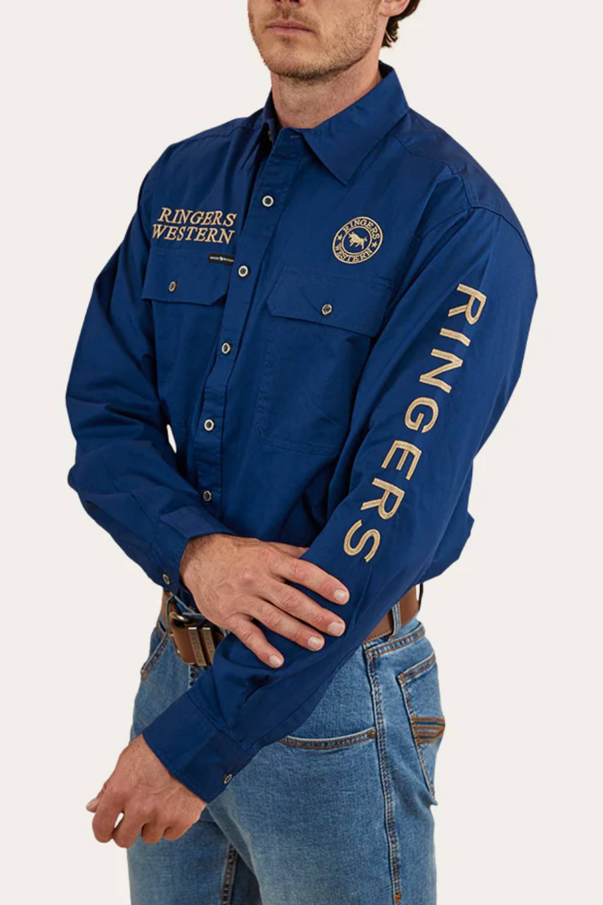A man wears the Ringers Western Hawkeye Embroidered Shirt in blue with UPF50+ protection, signature chest embroidery, a circular logo, "Ringers" on the sleeve, paired with blue jeans and a brown belt.