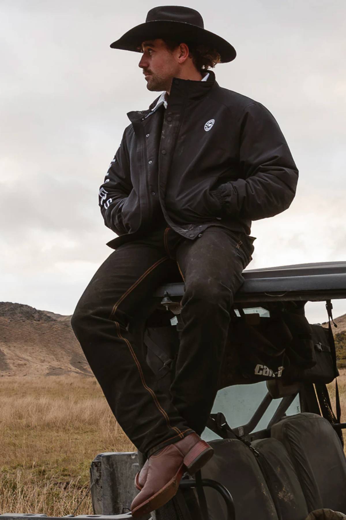 A man in a black cowboy hat, dark jeans, and a Ringers Western Kidman Jacket sits on a vehicle roof in an open grassy field with hills behind him, looking to the side with a relaxed posture.