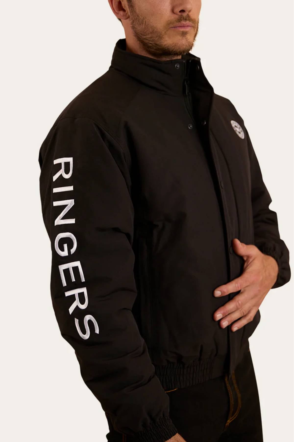 A man wears the Ringers Western Kidman Jacket, a black high-collared piece with "RINGERS" in bold white letters down the sleeve and a quilted lining. He has one hand in his pocket and faces slightly away from the camera.