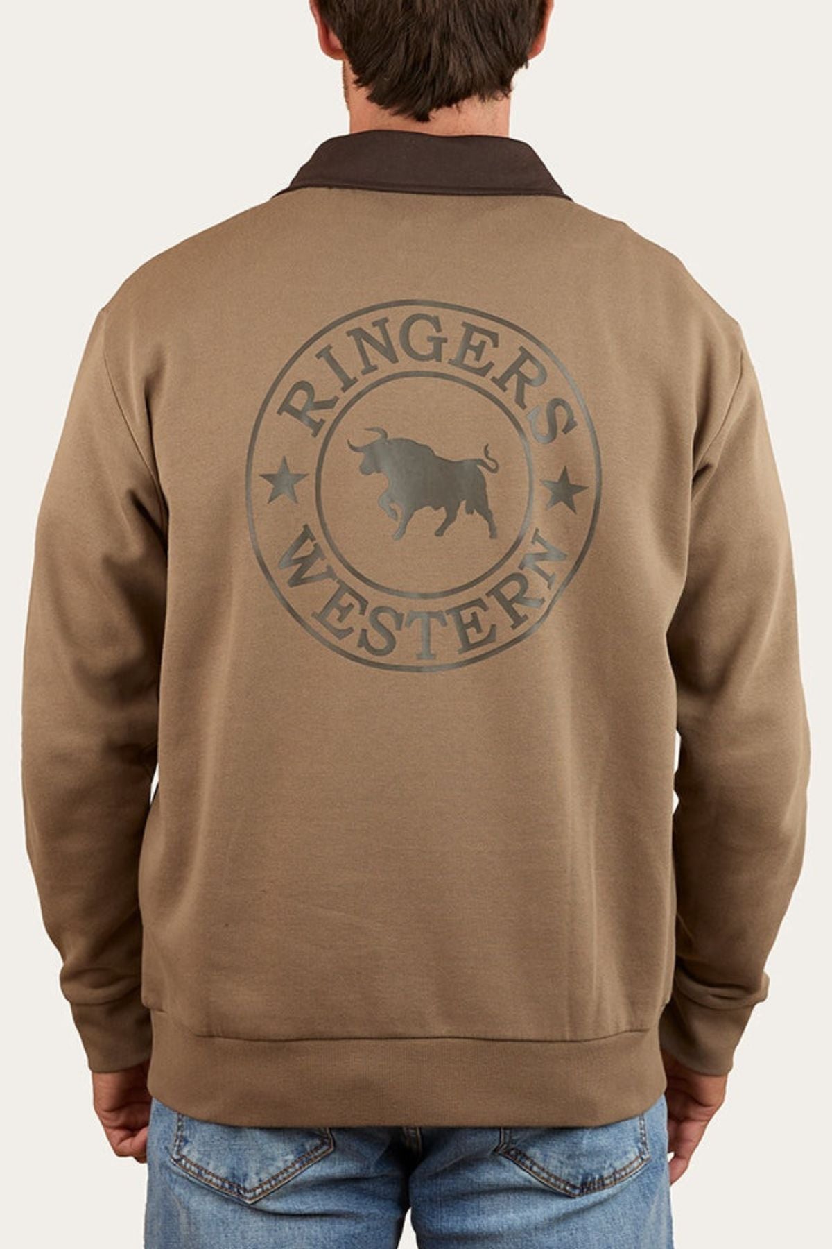 A man in blue jeans stands with his back to the camera, wearing a brown Ringers Western Lachlan 1/4 Zip Crew made from brushed fleece with the "Ringers Western" logo and a bull graphic on the back.
