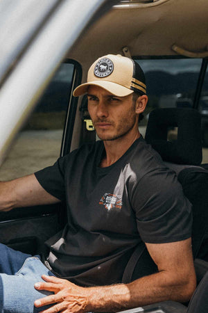 A man with a serious expression sits in a car, wearing a tan and black Ringers Western McCoy Trucker Cap with an adjustable strap, a black short-sleeve shirt, and blue jeans. He is positioned in the driver's seat, with his arm resting on the car's window frame.