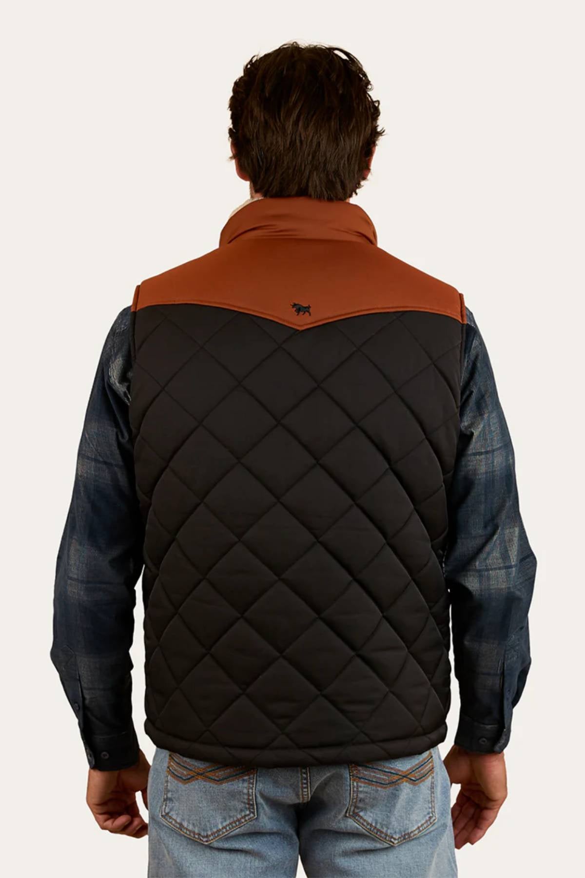 A man faces away, wearing the Ringers Western McKinlay Puffer Vest with a brown yoke over a blue plaid shirt and jeans. A small animal logo is displayed near the top center of the back, making it ideal for outdoor enthusiasts.