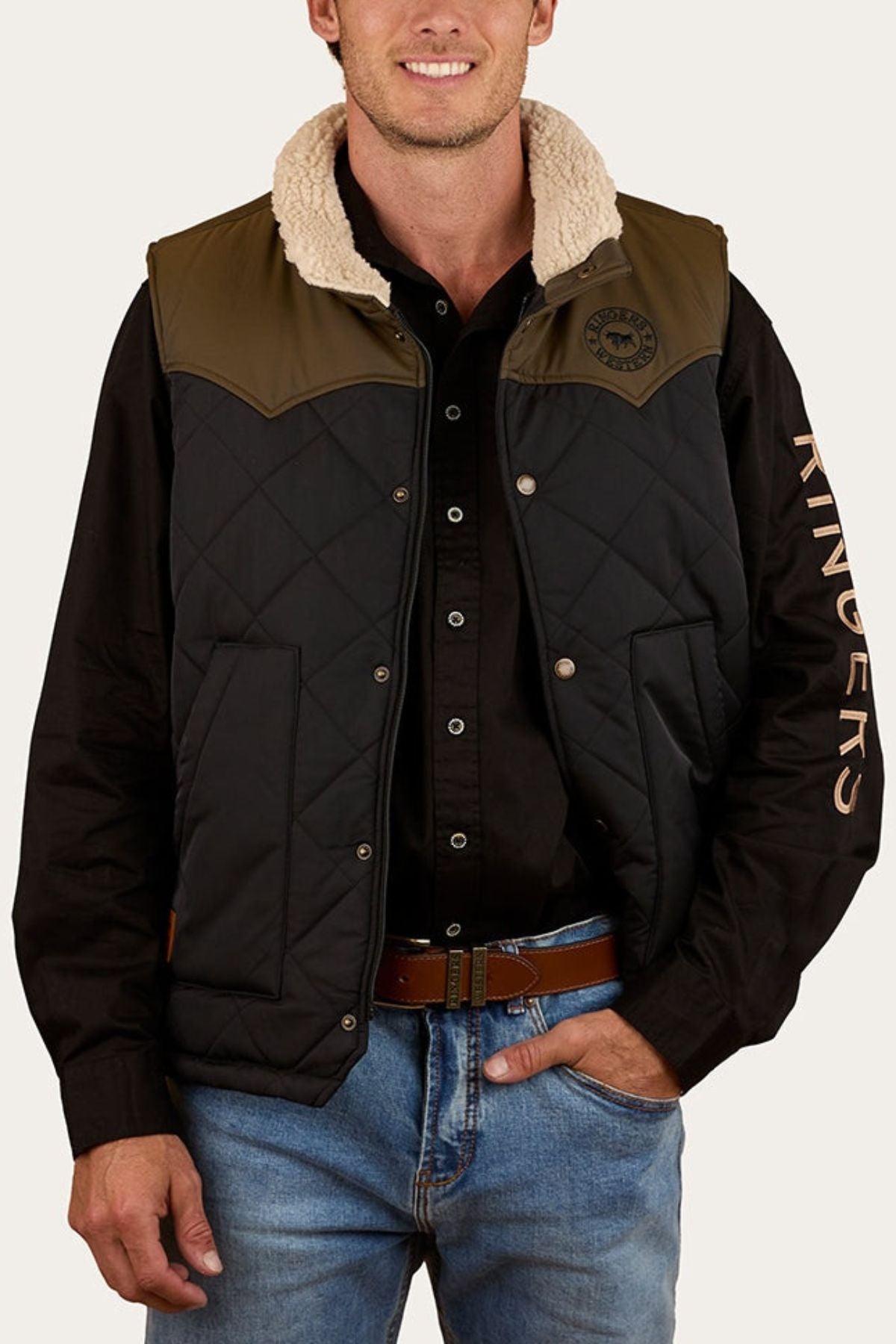 A man smiles with one hand in his pocket, wearing the Ringers Western McKinlay Puffer Vest with a sherpa collar over a black shirt featuring "RINGERS" on the sleeve and blue jeans—a casual outdoor style.