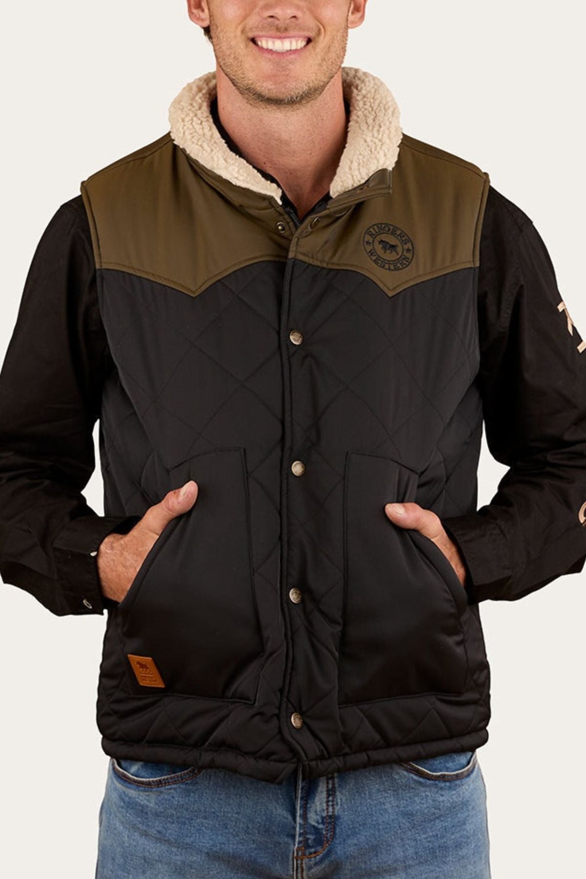 A man wears the Ringers Western McKinlay Puffer Vest, featuring a sherpa collar, snap buttons, and front pockets, over a black shirt and blue jeans. He smiles with his hands in the vest pockets.