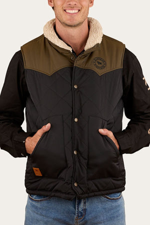 A man wears the Ringers Western McKinlay Puffer Vest, featuring a sherpa collar, snap buttons, and front pockets, over a black shirt and blue jeans. He smiles with his hands in the vest pockets.