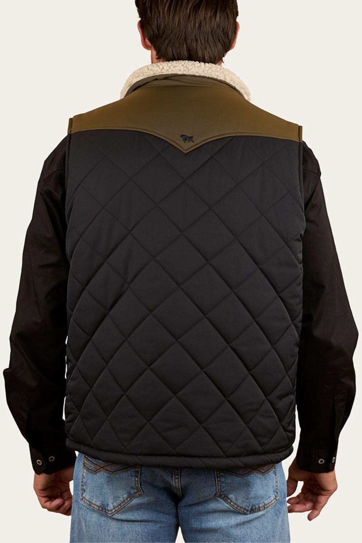 A person wears the Ringers Western McKinlay Puffer Vest with a black quilted design, olive green shoulder yoke, and subtle stitched pattern over a black long-sleeve shirt and blue jeans. The vest features the logo below the collar.
