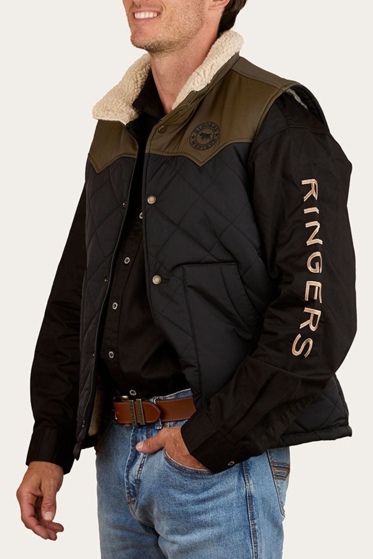 A man wears the Ringers Western McKinlay Puffer Vest—a black quilted vest with tan shoulders and a shearling collar, "RINGERS" on the sleeve. He smiles, left hand in his jeans pocket, facing slightly right.