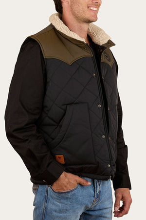 A man smiles and poses with one hand in his pocket, wearing a black Ringers Western McKinlay Puffer Vest with tan shoulder accents and a fleece collar over a black shirt and blue jeans.