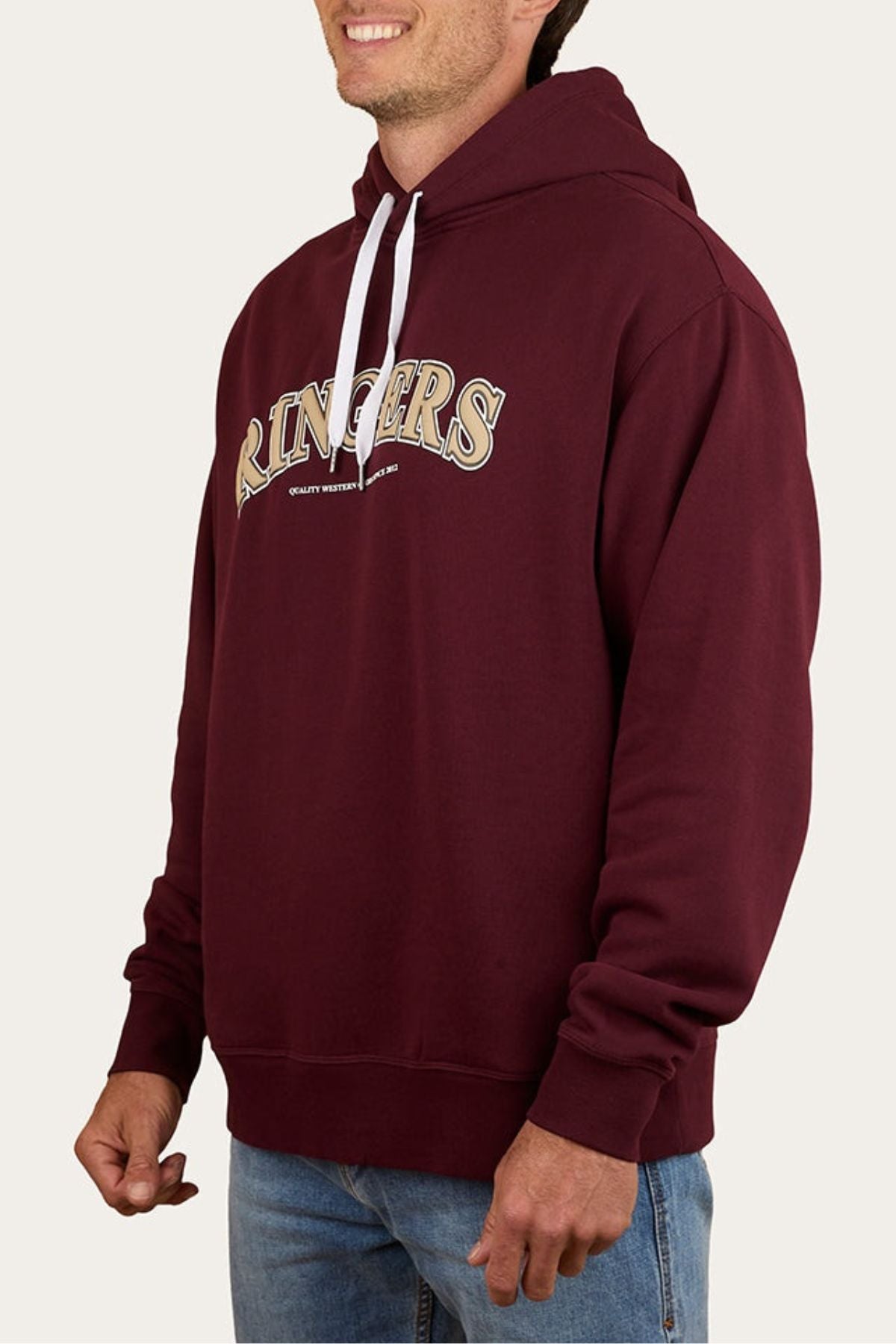 Someone stands against a plain background wearing the oversized maroon Ringers Western Princeton Hoodie, crafted from soft brushed fleece with a bold "RINGERS" print on the front, paired with light blue jeans.