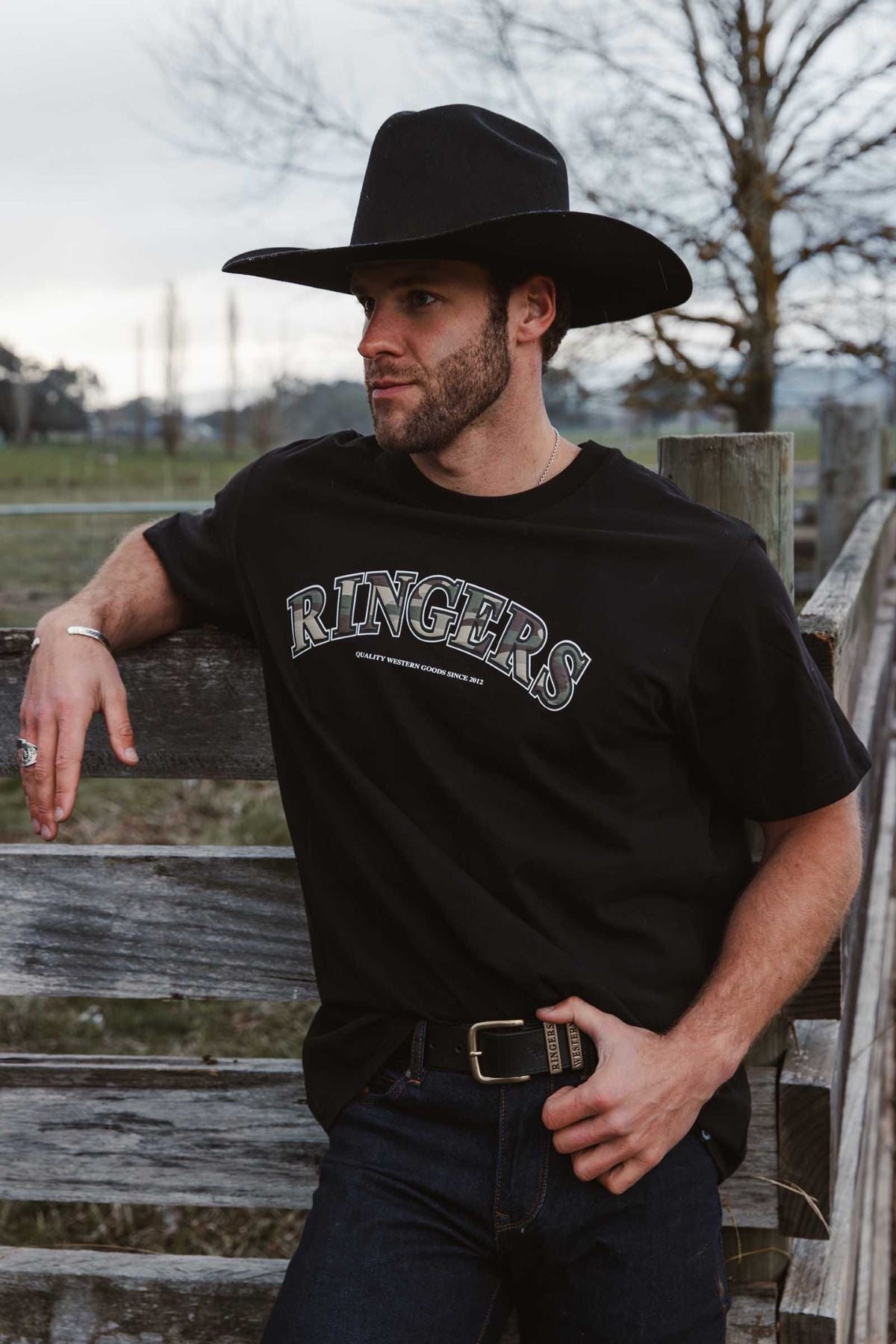 A man in a black cowboy hat and a black Ringers Western Princeton Loose Fit T-Shirt leans on a wooden fence outdoors, surrounded by trees and fields. His 100% cotton, loose fit shirt adds comfort to the relaxed country scene.