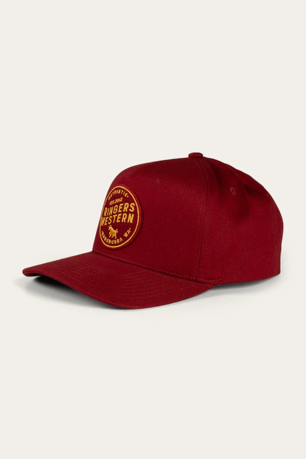A maroon Ringers Western Rye Baseball Cap, adorned with a yellow circular patch that reads "Ringers Western," is shown at an angle to highlight its front and side. The cap features an adjustable strap, and the off-white background accentuates the cap beautifully.