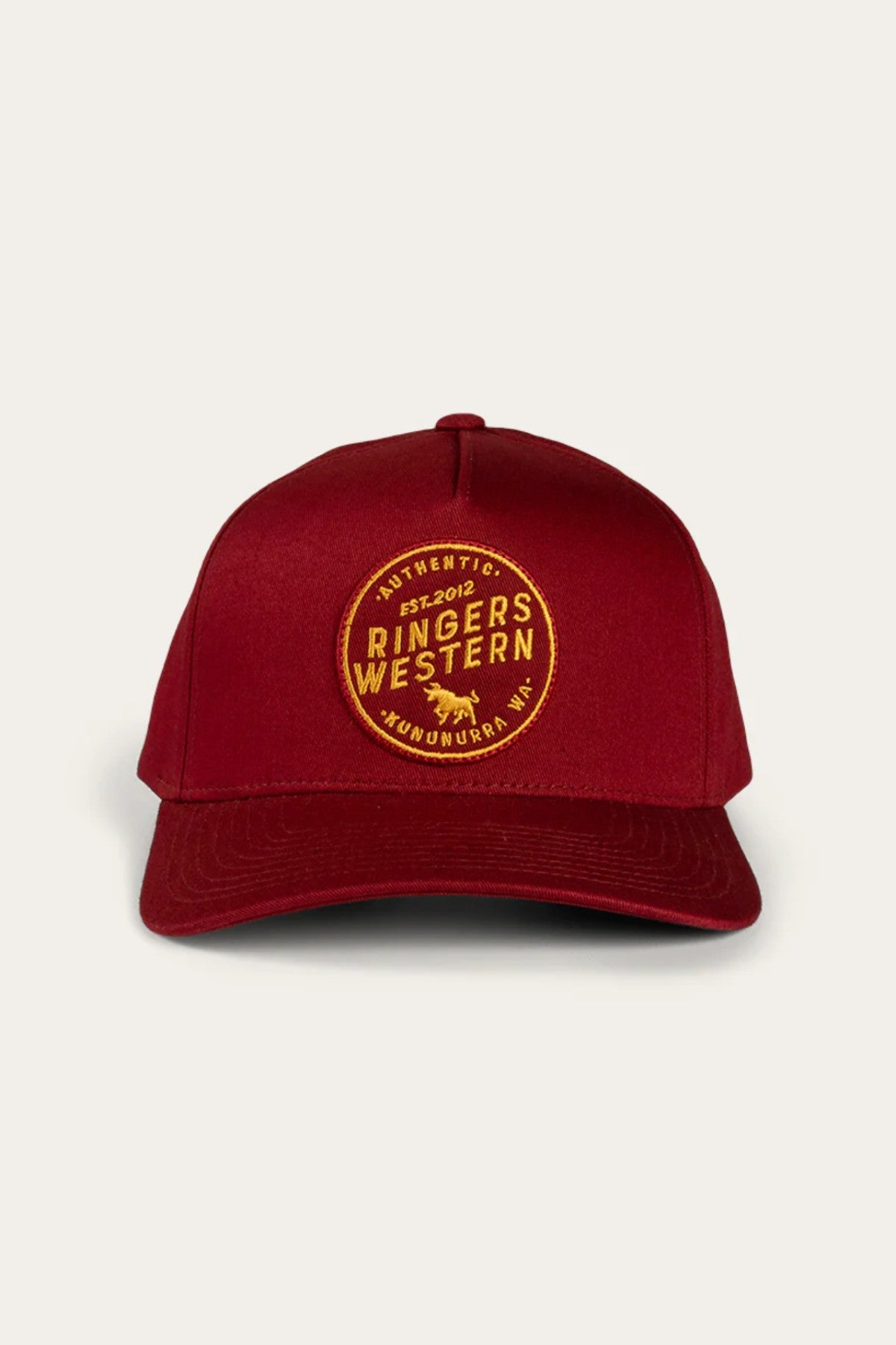 A front-facing image of the Ringers Western Rye Baseball Cap with a curved brim showcases its red design. The cap features a round, yellow embroidered patch on the front that reads "Authentic Ringers Western" and "Est. 2012, Wandoan, QLD," with a small bull icon centered within the patch. An adjustable strap at the back ensures a perfect fit.