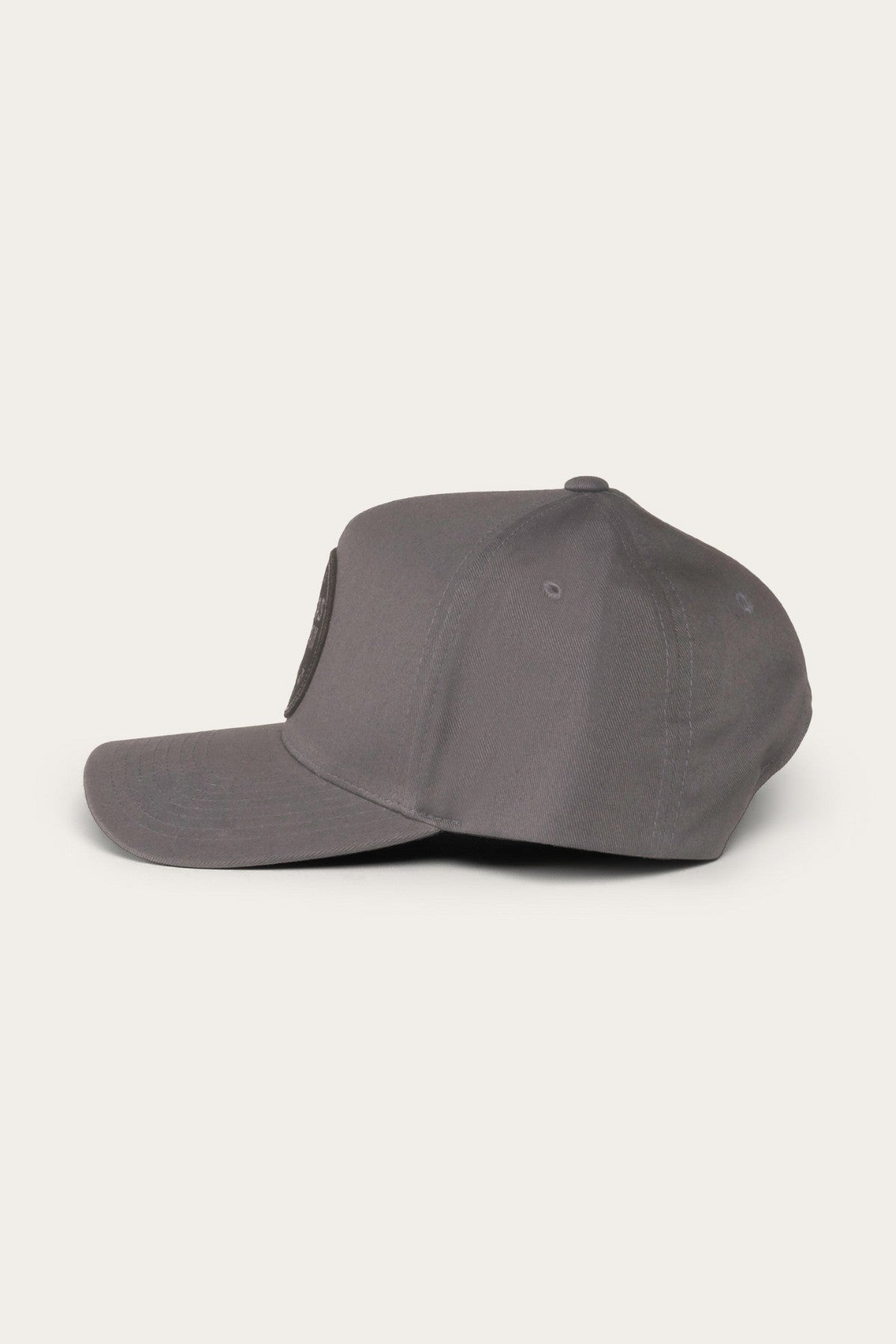 Side view of the Ringers Western Rye Baseball Cap in gray with a flat brim, six-panel construction, and stitched ventilation eyelets. The Cotton Twill cap features a circular patch logo on the front panel and is set against a plain, light background.