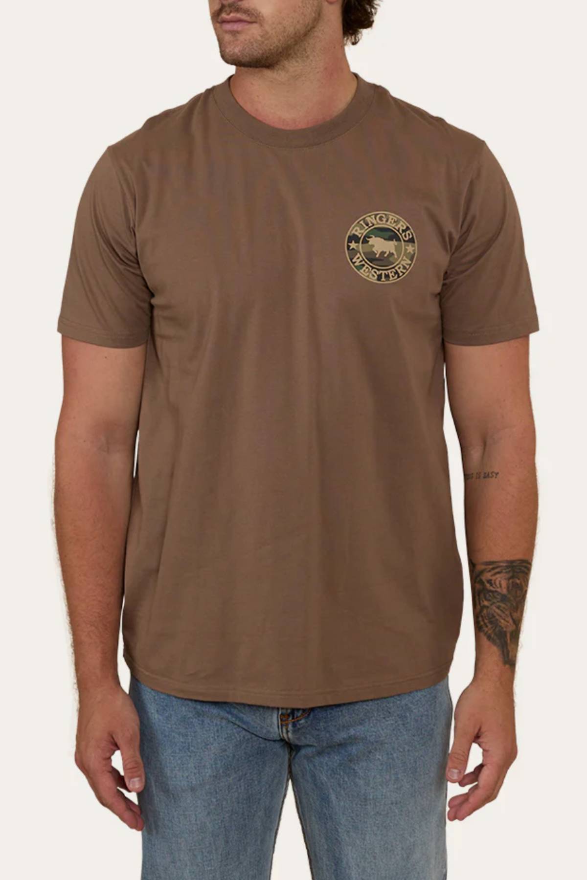 A man wears a brown Ringers Western Signature Bull Loose Fit T-Shirt, made from 100% cotton jersey with a small circular logo on the left chest, paired with blue jeans. The relaxed tee reveals tattoos on his right forearm.