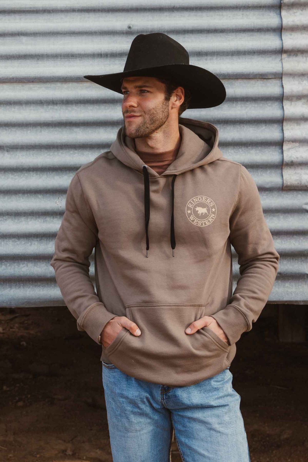A man wearing a black cowboy hat, blue jeans, and a Ringers Western Signature Bull Pullover Hoodie with front pocket stands in front of a corrugated metal wall, looking to the side with his hands in his hoodie pocket.