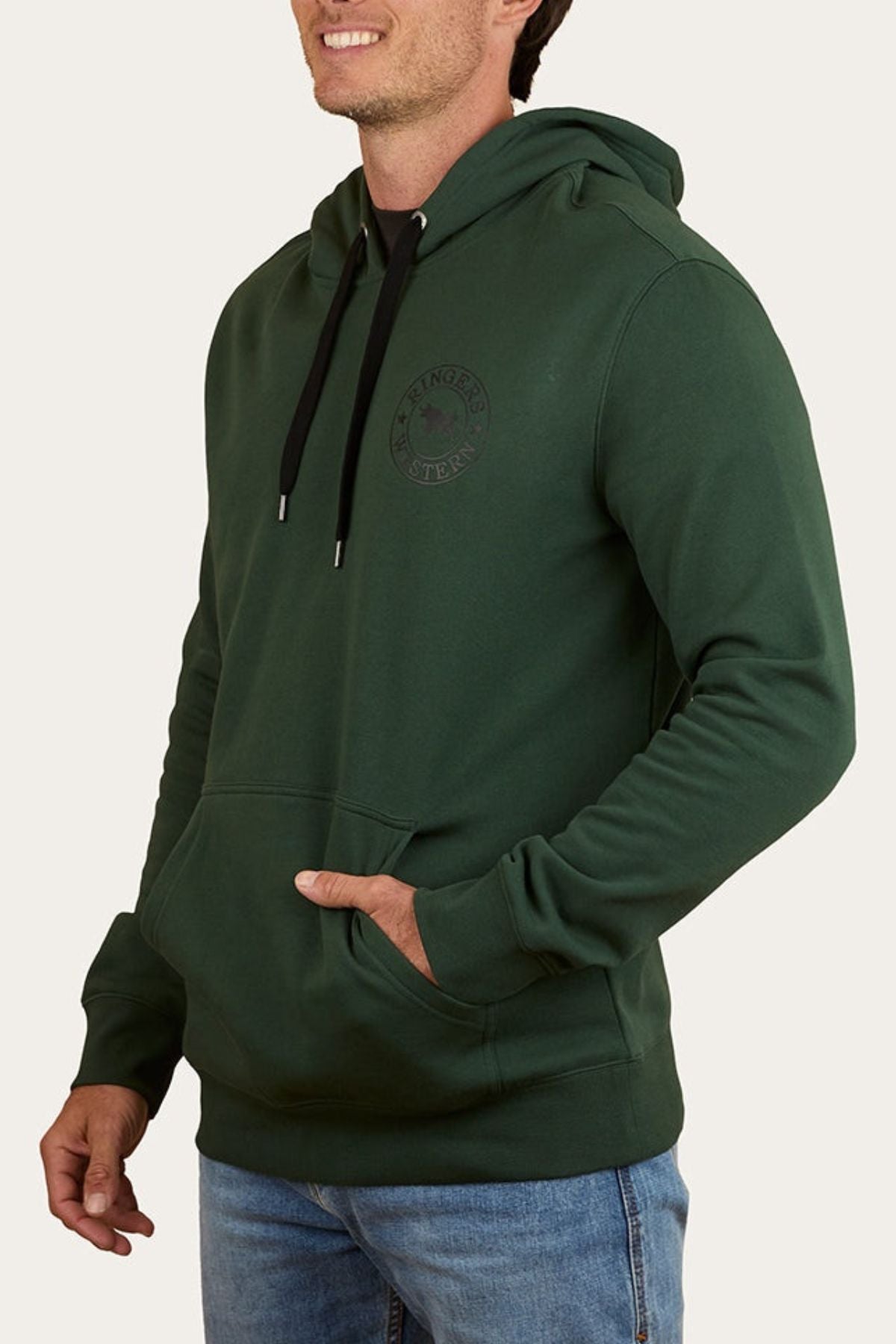 Wearing a dark green Ringers Western Signature Bull Pullover Hoodie with a front pocket and black drawstrings, paired with light blue jeans, a smiling person stands with one hand in their hoodie pocket.