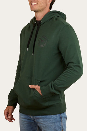 Wearing a dark green Ringers Western Signature Bull Pullover Hoodie with a front pocket and black drawstrings, paired with light blue jeans, a smiling person stands with one hand in their hoodie pocket.