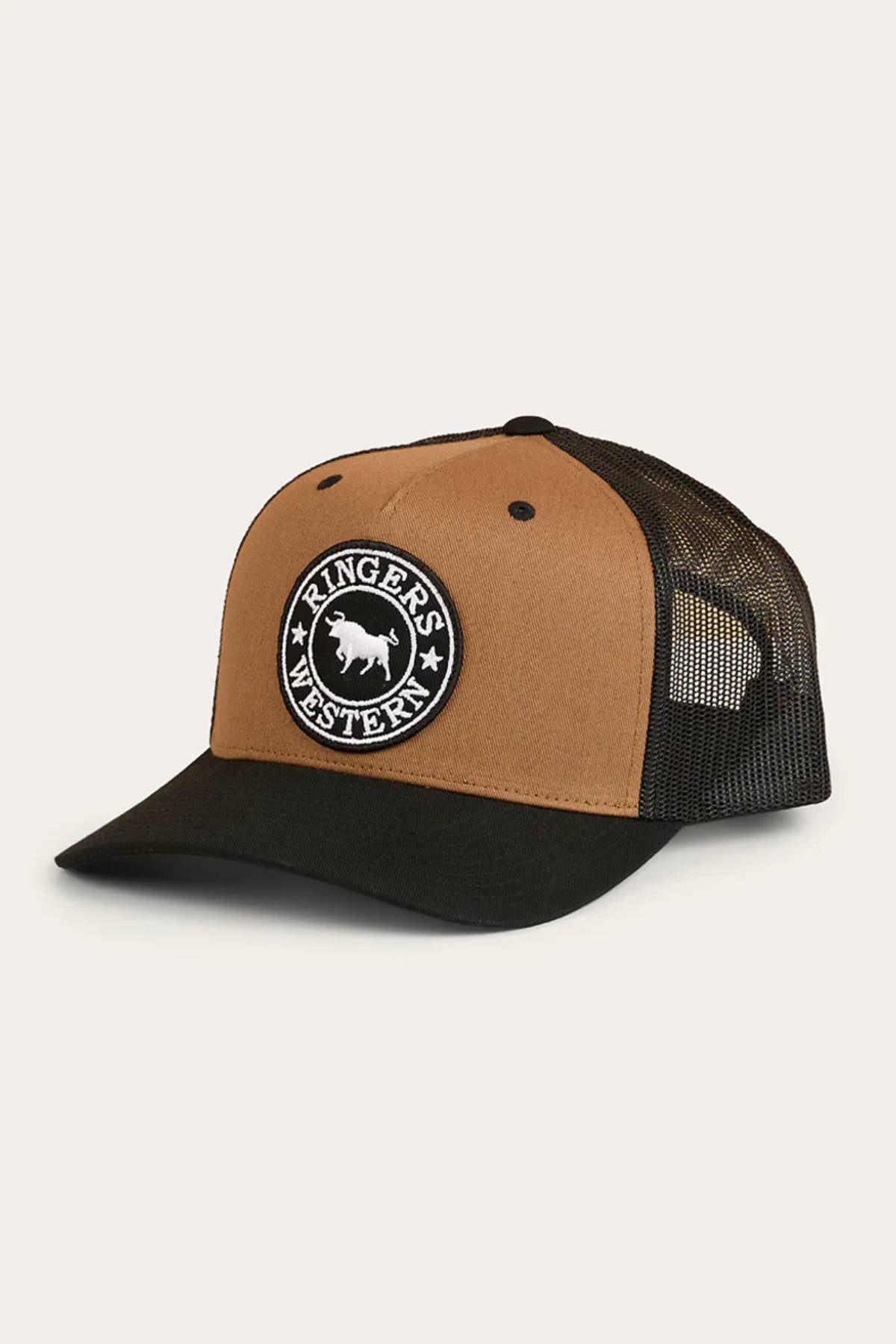 The Ringers Western Signature Bull Trucker Cap is tan and black with a black mesh back, adjustable strap, and features a circular "Ringers Western" bull patch on the front panel.