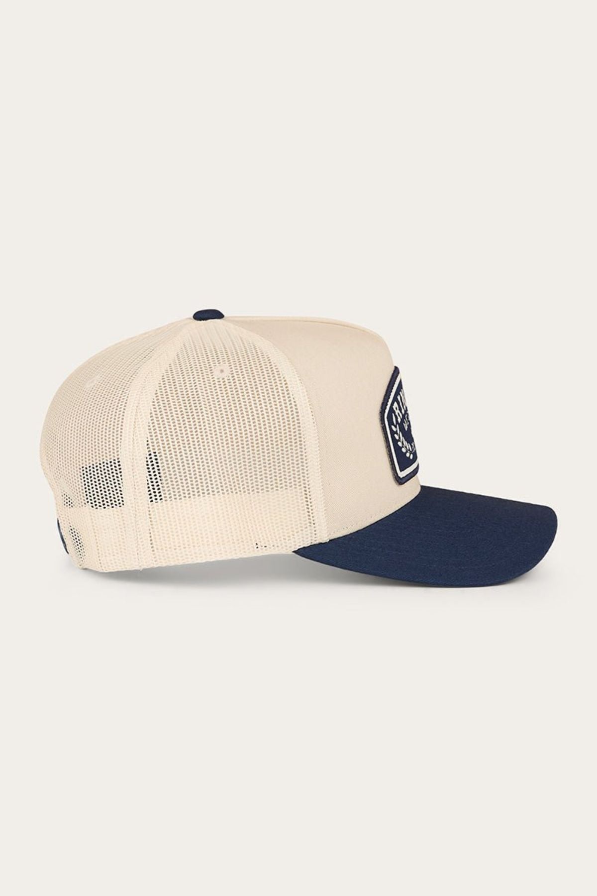 The Ringers Western Wheatbelt Trucker Cap is a cream and navy cap featuring a breathable mesh back and a flat brim, shown from the side. The front is adorned with a rectangular patch logo, and it comes with an adjustable snapback for a comfortable fit.