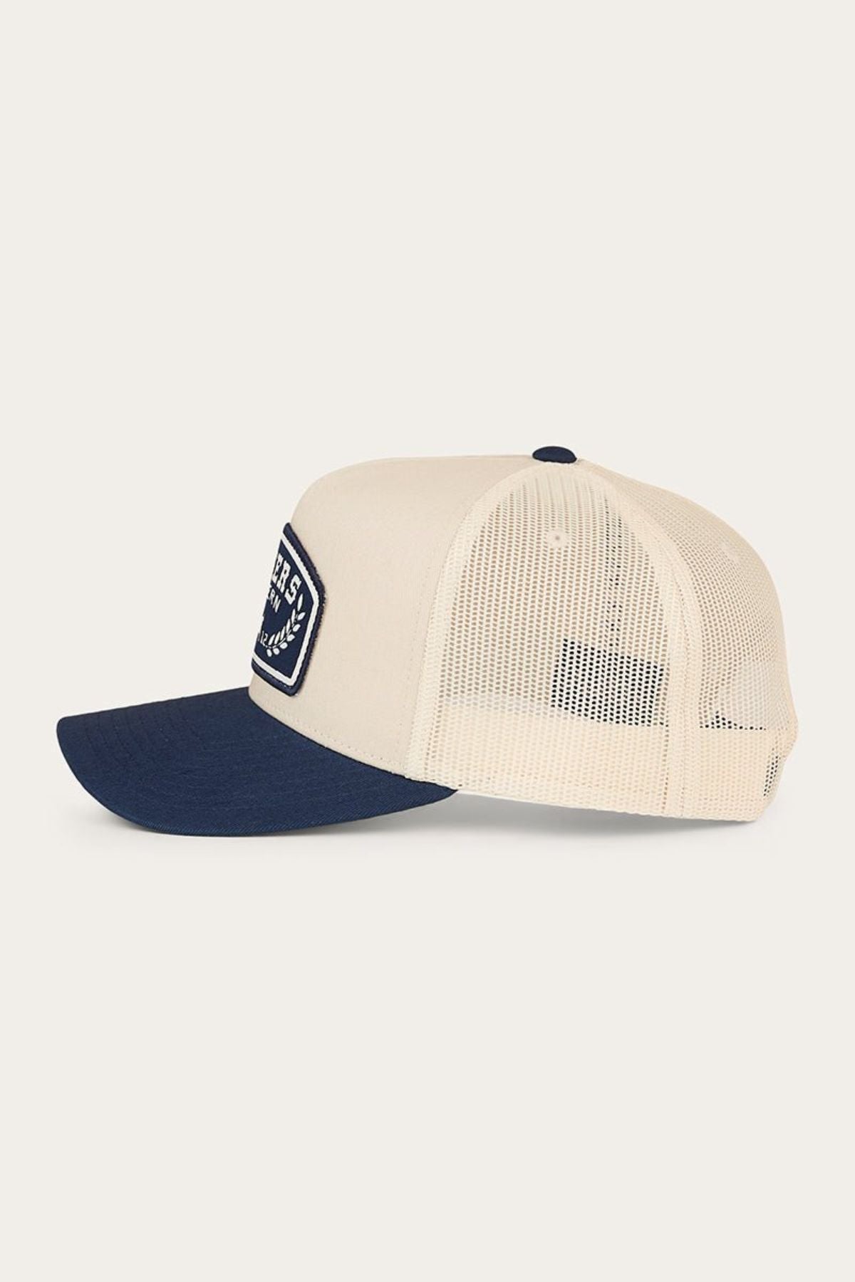 The Ringers Western Wheatbelt Trucker Cap in beige is displayed in a side view against a plain background, featuring a navy blue bill and a rectangular patch on the front left panel. This mid-profile cap includes an adjustable snapback closure for comfortable all-day wear.