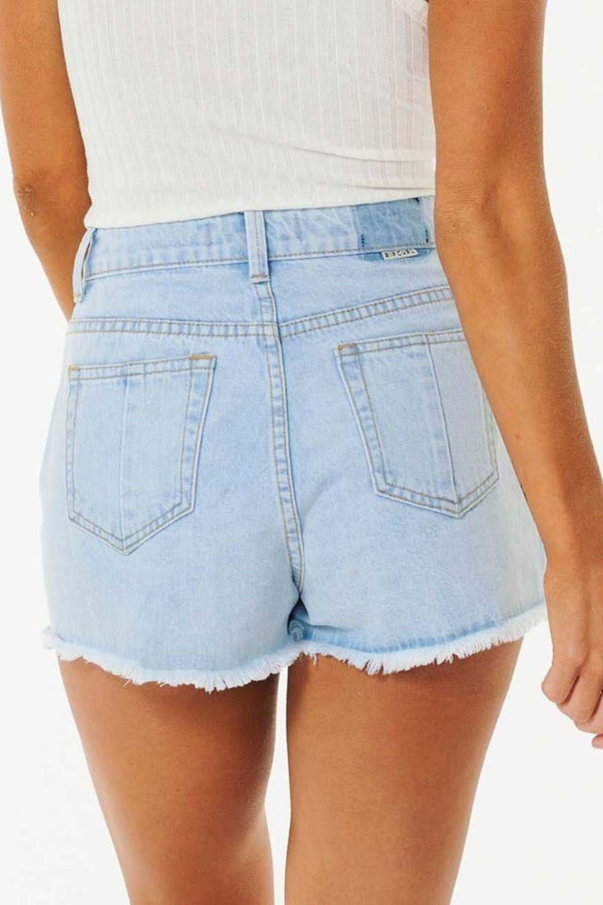A person is shown from the back wearing the "Rip Curl Amy High Waist Short" in light blue denim, featuring a high waist and frayed hems, paired with a sleeveless white top against a plain white background that subtly evokes summer adventures.