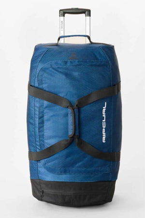The Rip Curl Jupiter Search Ref 80L Travel Bag is a large blue duffel with a retractable handle, black straps, and a vertical Rip Curl logo. This water resistant rolling bag stands upright on a plain white background.