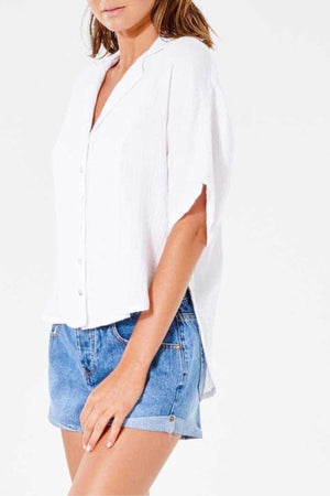 A person in a relaxed-fit, white short-sleeve Rip Curl Premium Surf Shirt and denim shorts stands against a plain white background. The image is cropped just above the shoulders, leaving the head out of frame, highlighting the casual vibe.