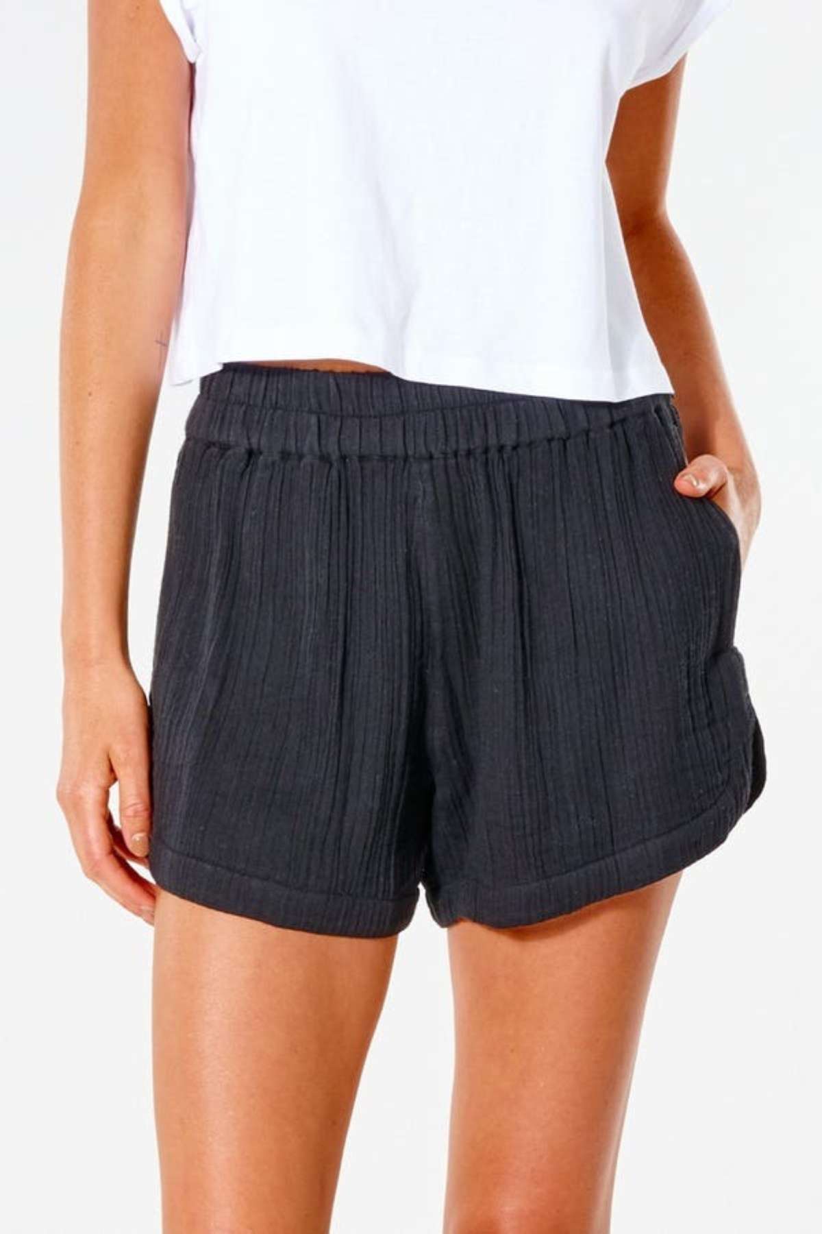 A surf enthusiast wearing a white cropped shirt stands with one hand in their pocket against a plain background, showcasing the high-waisted Rip Curl Premium Surf Short.
